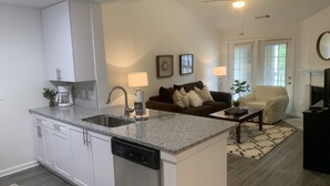 A modern kitchen with white cabinets, a dishwasher, and a granite countertop. There is a dining area with a brown couch and a coffee table. - Fabulous Fully-Furnished Apartment in Alpharetta (Alpharetta)