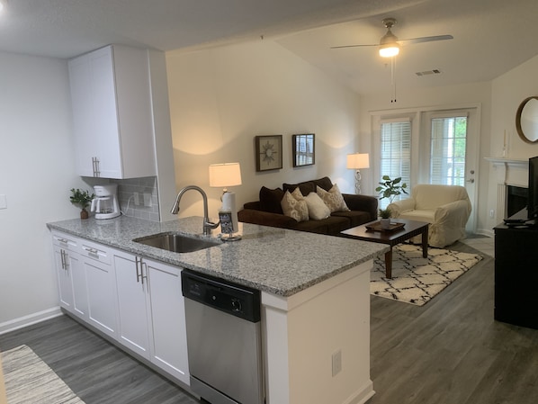 A modern kitchen with white cabinets, a dishwasher, and a granite countertop. There is a dining area with a brown couch and a coffee table.