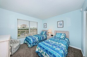 A bedroom with two beds, a nightstand, a lamp, a chair, and a window with blinds.