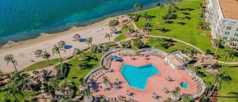 An aerial view of a resort with a central swimming pool, a beach, and palm trees.