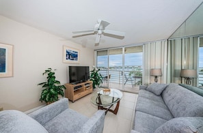 A modern living room with a flat-screen TV, two potted plants, a glass coffee table, and a grey sofa.