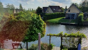 A lakeside view with a patio area, a pergola, and a row of houses in the background. - Luxury Holiday Home, Large Garden, Free Motorboat Usage for Our Guests (Medemblik)