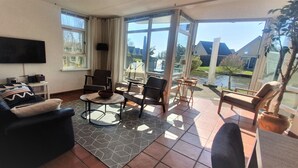 A living room with a sofa, armchairs, a coffee table, and a TV. There are large windows with a view of a house and a body of water. - Luxury Holiday Home, Large Garden, Free Motorboat Usage for Our Guests (Medemblik)