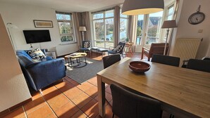 A living room with a blue sofa, a wooden dining table, and a large window with curtains. - Luxury Holiday Home, Large Garden, Free Motorboat Usage for Our Guests (Medemblik)