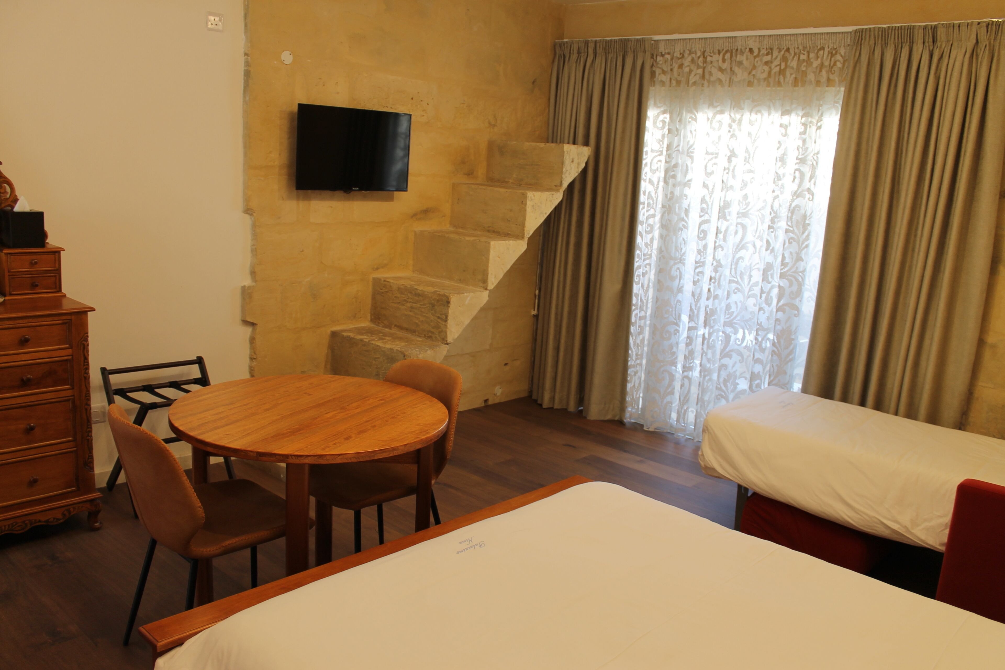 A room with a wooden dining table, chairs, a bed, a TV mounted on the wall, and a staircase.