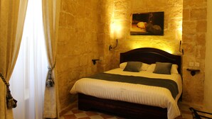 A bedroom with a stone wall, a bed with white bedding, a painting above the bed, and a wooden headboard. - Palazzino Nina Boutique Hotel (Qormi)