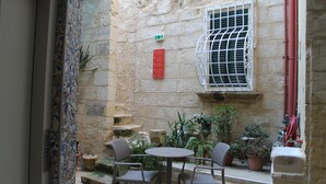A stone building with a small patio area featuring a round table and two chairs, surrounded by potted plants and a window with metal bars. - Palazzino Nina Boutique Hotel (Qormi)