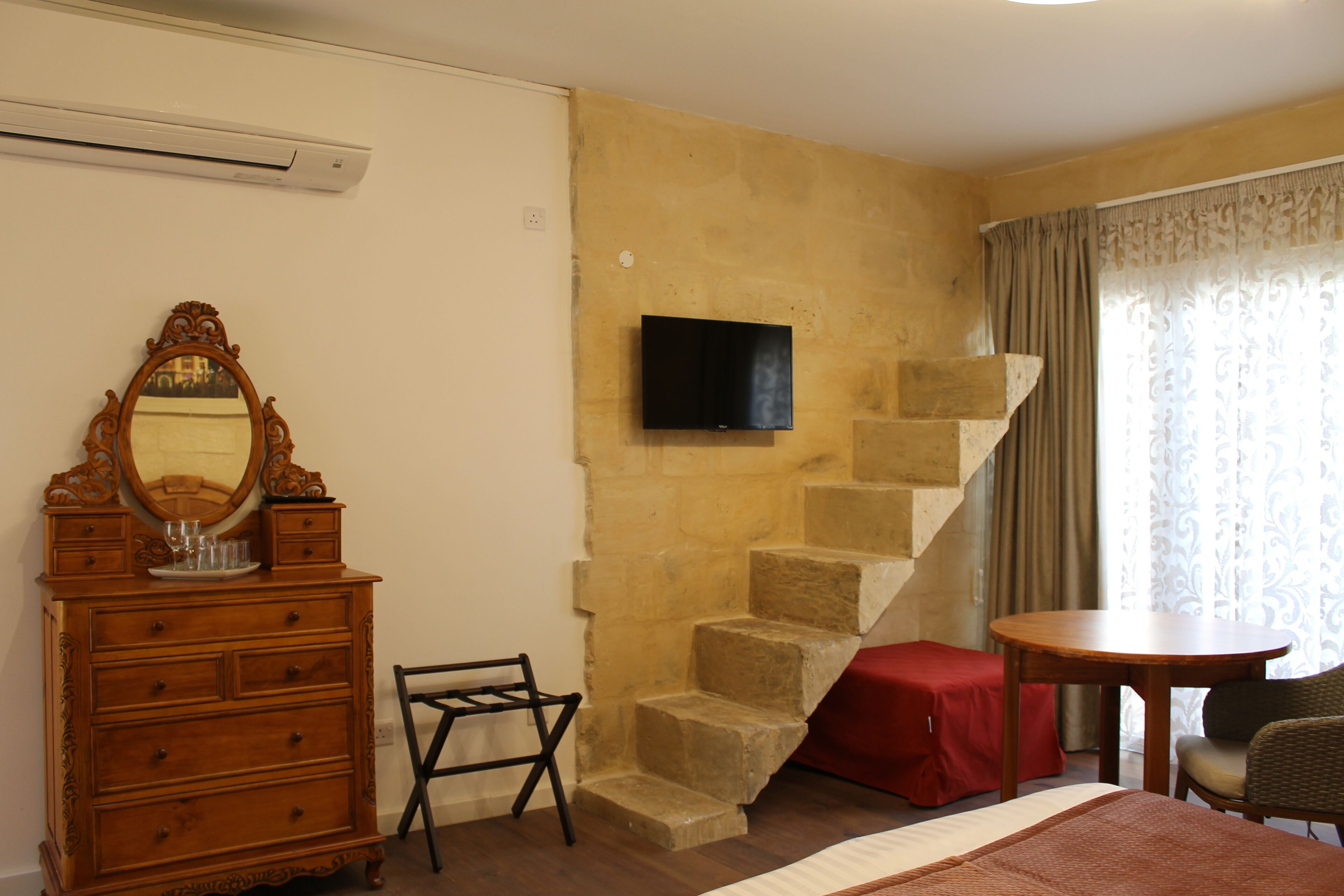 Une chambre avec une commode en bois, un miroir, un téléviseur fixé au mur, un escalier, une petite table avec une chaise et une fenêtre avec des rideaux.