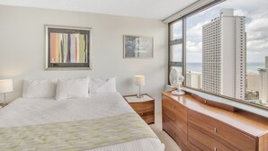 A hotel room with a large bed, a dresser, a nightstand, and a window with a city view. - Steps to the Beach! Gorgeous Ocean-View Condo with Complimentary Parking (Honolulu)