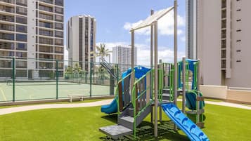 Ein Spielplatz mit Rutschen und Klettergerüsten, umgeben von einem Tennisplatz und Hochhäusern.