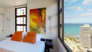 Habitación de hotel con una cama grande, almohadas naranjas, un cuadro abstracto de colores, una mesita de noche, una lámpara y vistas al océano y edificios.