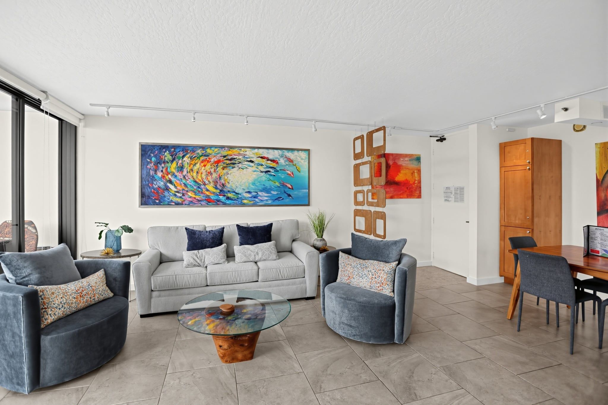 A modern living room with a grey sofa, two armchairs, a glass coffee table, and a dining area with a wooden cabinet and table.