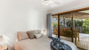 A bedroom with a bed, a ceiling fan, a nightstand, and a view of a balcony with a wooden deck and greenery. - Sunshine Beach Shack - Pet Friendly close to the beach (Sunshine Beach)