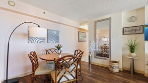 A dining area with a round table, four chairs, and a floor lamp. There is a framed picture on the wall and a small potted plant on a side table.