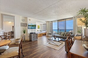 A spacious living area with a dining table, chairs, a sofa, and a large window offering a view of the outdoors. - Beautifully Renovated 32nd Floor Deluxe Ocean View Waikiki Condo (Honolulu)