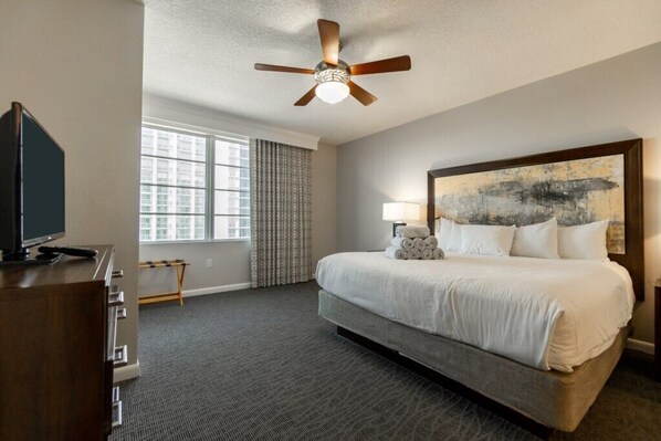 A hotel room with a bed, a television, a ceiling fan, a window with curtains, and a painting on the wall.
