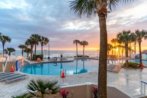 A pool area with palm trees, a curved pool, and a view of the sunset over the ocean.
