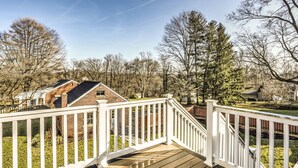 Eine Holzterrasse mit weißen Geländern, umgeben von Bäumen und einem Ziegelhaus im Hintergrund.
