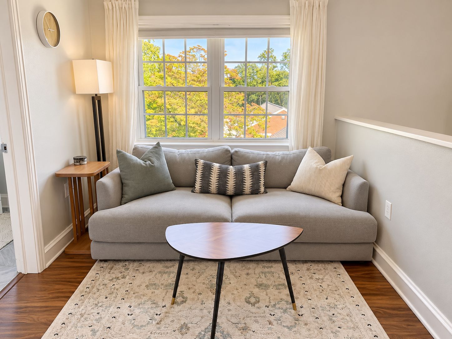 A living room with a sofa, a small table, a lamp, and a window with a view of trees.