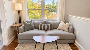 A living room with a sofa, a small table, a lamp, and a window with a view of trees.
