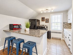 A kitchen with a granite countertop, a red microwave, a black refrigerator, and a stove. There are three blue stools and a potted plant on the counter.