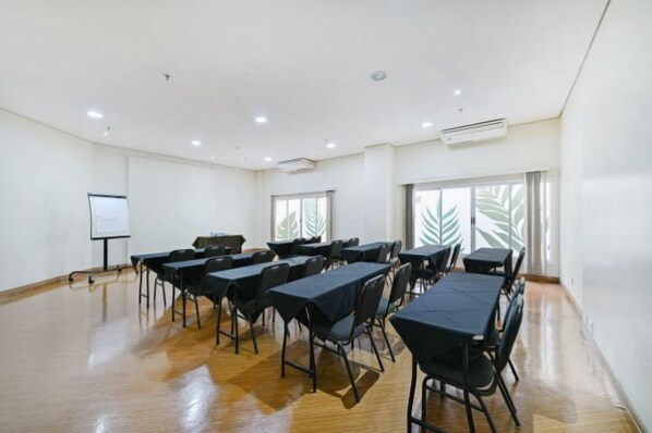 Un salón de conferencias con mesas y sillas, una pizarra y ventanas grandes con un diseño de hojas.