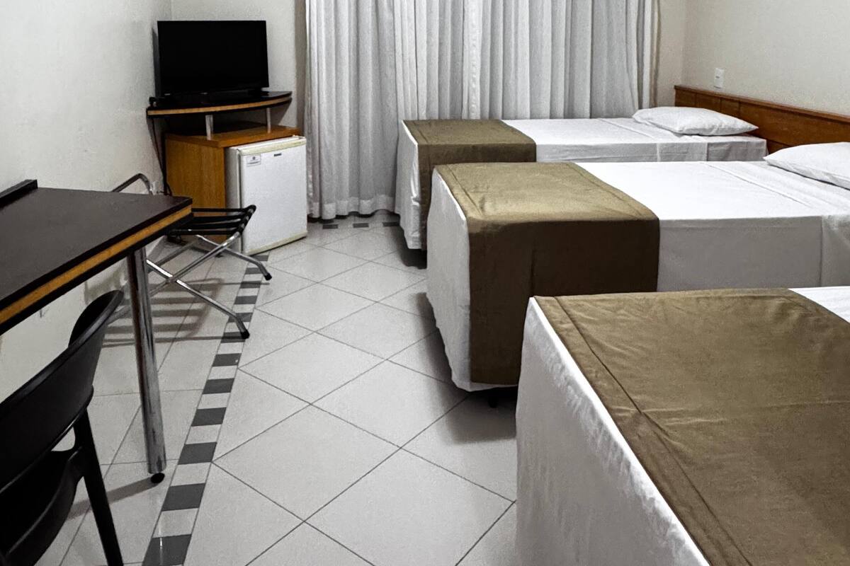 A hotel room with a desk, two single beds, a small kitchenette, and a large window with curtains.