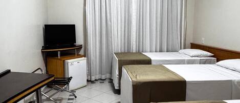 A hotel room with a desk, two single beds, a small kitchenette, and a large window with curtains.