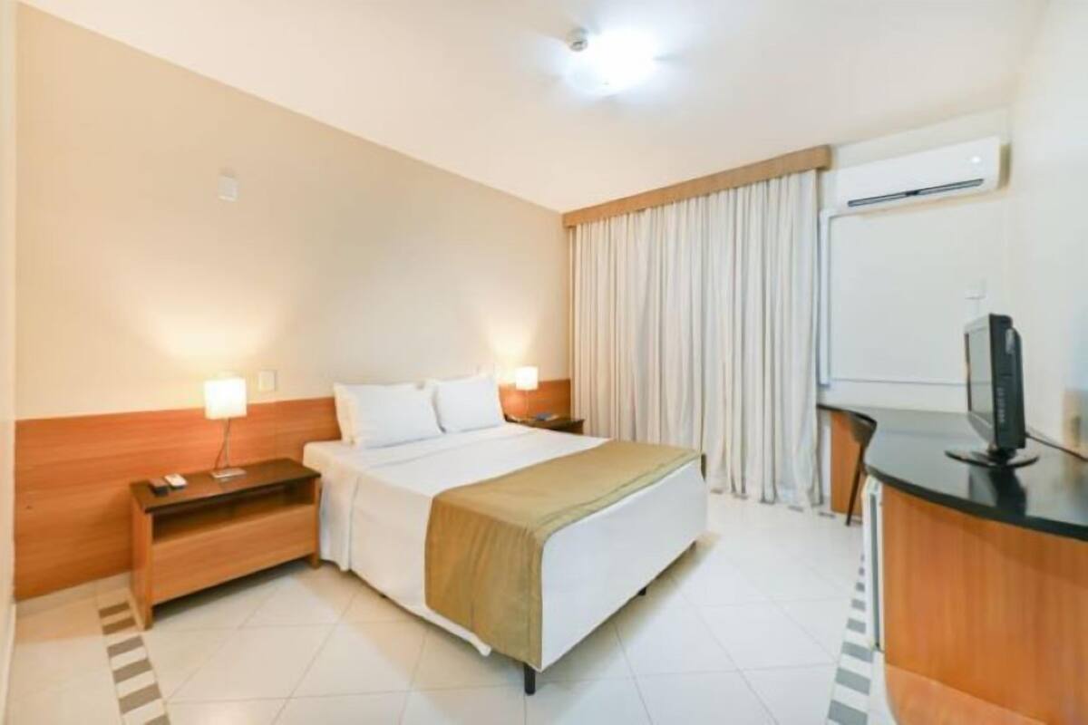 A hotel room with a bed, bedside tables, a desk, a TV, and an air conditioning unit.