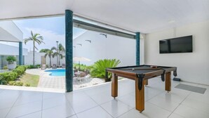 A modern indoor pool area with a pool table, a TV mounted on the wall, and a view of the outdoor pool and patio furniture. - Hotel Nacional Inn Cuiabá (Cuiabá)
