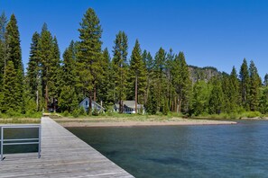 A wooden dock extending into a calm lake, surrounded by dense green forest and a few houses.