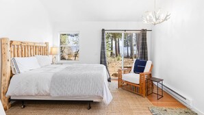 A bedroom with a large bed, a wooden headboard, a window with a view of trees, a wicker chair, and a chandelier. - Belleview Lakefront by Tahoe Getaways - Hot Tub, Pier, Buoy and Guest House (Homewood)