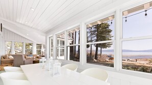 Une pièce spacieuse avec une grande table à manger, des chaises blanches et de grandes fenêtres donnant sur un lac et des montagnes.