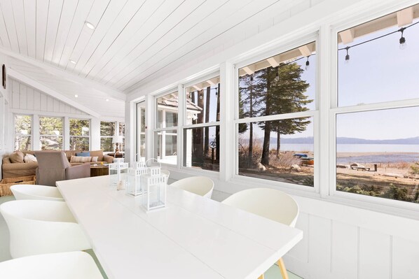 A spacious room with a large dining table, white chairs, and large windows offering a view of a lake and mountains.