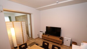 Ett vardagsrum med ett matbord i trä, stolar, en platt-TV, ett litet kylskåp och en traditionell japansk skärm.