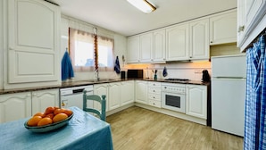 A compact kitchen with white cabinets, a stove, a microwave, and a refrigerator. A table with a bowl of fruit is visible in the foreground.