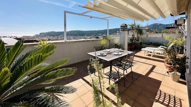 A rooftop patio with a table set for dining, surrounded by potted plants and a view of the city and hills.