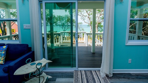 A room with a sliding glass door leading to a deck with a view of the ocean. There is a blue sofa with a patterned pillow and a small table with a surfboard. - Fish Fever, Russell Island Bahamas (Russell Island / Spanish Wells)