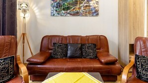 Een bruine leren bank met gekleurde kussens, een vloerlamp en een kleurrijk abstract schilderij aan de muur.