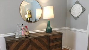 Une commode en bois à motifs en chevrons, sur laquelle est posée une barquette de bouteilles, un miroir rond et une lampe de table verte, le tout adossé à un mur orné d’une assiette décorative.