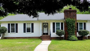 Une maison de plain-pied avec une cheminée en briques, une porte d’entrée et un petit jardin devant, avec un chemin menant à la porte.
