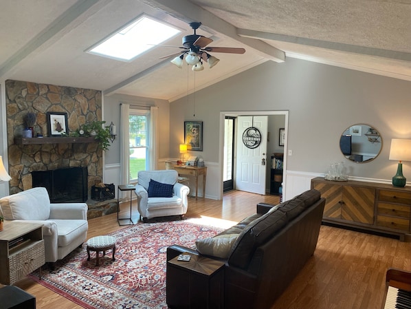 A spacious living room with a stone fireplace, a ceiling fan, a large window, and a cozy seating area with a patterned rug.