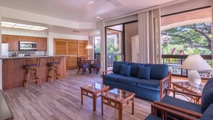 Una sala de estar moderna con un sofá, una mesa de centro y sillas. Hay una zona de cocina con armarios de madera y un microondas. La habitación tiene ventanas grandes con vistas a árboles.