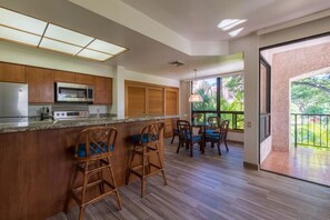 A modern kitchen with wooden cabinets, a stainless steel refrigerator, and a built-in microwave. There are two wicker bar stools with blue cushions. The kitchen has a large window providing a view of greenery outside. - Shores Waikoloa - Remodeled 1 Bedroom Condo (Waikoloa Village)