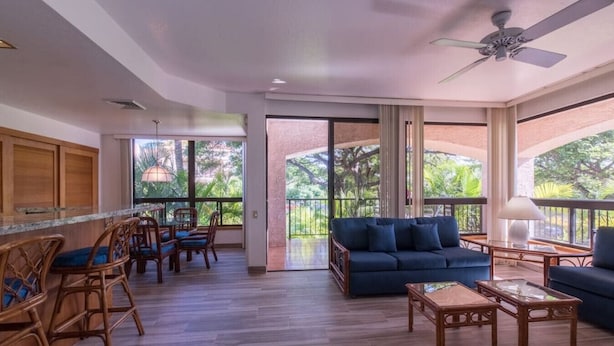 Amplia sala de estar con un sofá azul oscuro, mobiliario de madera y grandes ventanales que dan a vistas de vegetación.