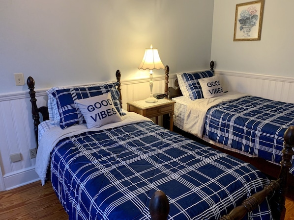 Une chambre avec deux lits, chacun recouvert d’une couette et de coussins à carreaux bleus et blancs, ornés du texte « BONNE AMBIANCE ». Une table de chevet avec une lampe se trouve entre les deux lits.