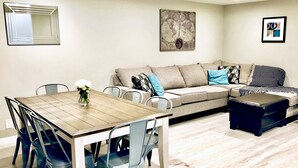 A dining area with a wooden table and blue chairs, a beige sofa with patterned cushions, and a black ottoman. - Family-Friendly, Renovated Suite (Extra Cleaning) (Orem)