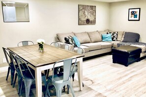 A dining area with a wooden table and blue chairs, a beige sofa with patterned cushions, and a black ottoman.