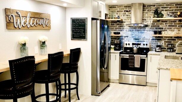 A modern kitchen with a stainless steel refrigerator, oven, and gas stove. There are black bar stools and a wooden countertop. A 'Welcome' sign and a chalkboard menu are on the wall. - Family-Friendly, Renovated Suite (Extra Cleaning) (Orem)