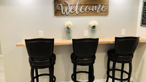 A kitchen area with a wooden bar and three black barstools. A 'Welcome' sign is mounted on the wall above the bar. - Family-Friendly, Renovated Suite (Extra Cleaning) (Orem)